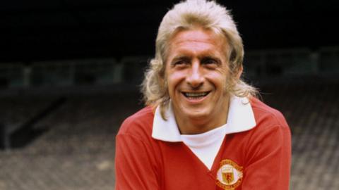 Denis Law, with a blonde mullet, in his early 1970s Manchester United red shirt with extended white V-necked collar