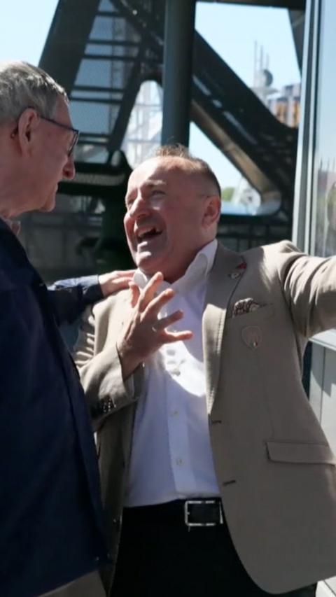 A man in a brown suit singing and holding his arms out in exclamation 