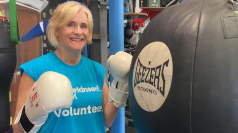 A woman wearing white boxing gloves and a blue t-shirt with the words "Parkinson's UK volunteer" stands next to a punch bag.