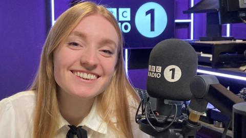 A lady with blonde hair in a white shirt looking directly at the cameral. A black BBC Radio 1 microphone is to her right. The walls of the studio are purple.