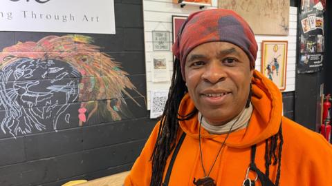 Roland Burt is looking at the camera. He is wearing an orange hoodie, elephant pendant and a red and navy bandana over dreadlocks. Behind him is a wall displaying a drawing of a lion and other artwork.