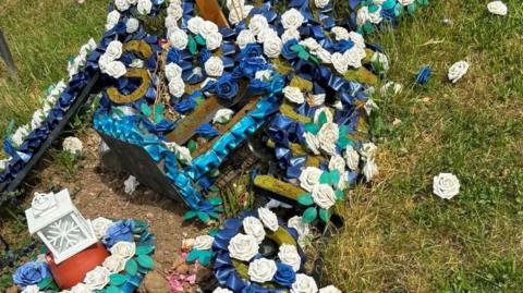 A close up of a grave with blue and white faux floral arrangements. Some of the flowers have fallen off and are lying on the grass.