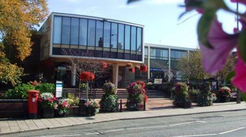 A municipal building on a street in a town, with manicured bushes and a red postbox outside. The building is two storey, with a large glass frontage on the second floor, which is supported by pillars. Steps lead up to a receded entrance.