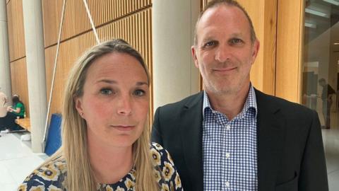 Sarah Hawkins, who has long blonde hair and is wearing a flowery top, stands to the left of her husband Jack. He has stubble and is wearing a blue gingham shirt and dark suit jacket.