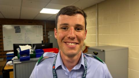 Bradley Lefley-Kemp is smiling and wearing a blue uniform with green lapel covers and a Poppy Appeal lanyard around his neck. He is standing in an office with desks and a filing cabinet in the background. 