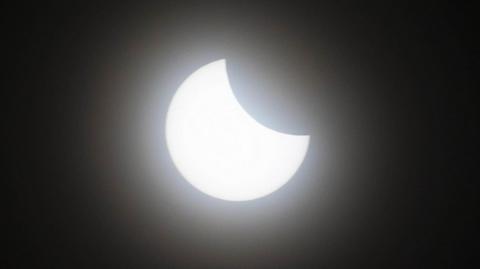 A picture of a partial eclipse showing a bright white sun with a crescent over its upper-left quarter. The background is black. 