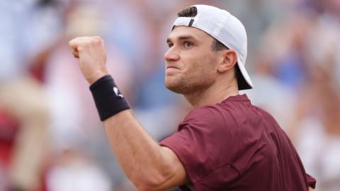 A man on a tennis court raising a clenched fist. He is wearing a baseball cap and maroon t-shirt.