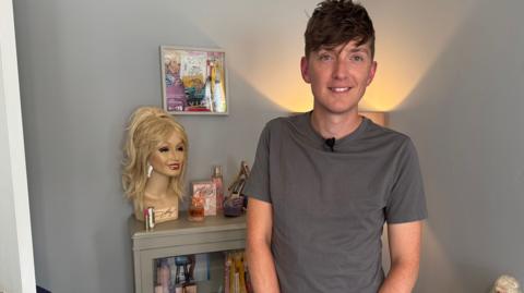 The photo shows a person standing in a room beside a display cabinet filled with Dolly Parton memorabilia, including CDs, photographs, and collectible items. On top of the cabinet is a mannequin-style wig head with a blonde wig, alongside perfume bottles, a replica high-heeled shoe and framed souvenirs.