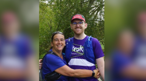 A woman smiles widely as she hugs a taller man who is wearing a gap and glasses and also smiling. They are outside and wearing purple tops.