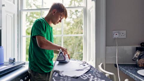 Stock photo shows a person ironing a T shirt in front of a window in their kitchen, next to a plug socket and with the washing machine behind them.