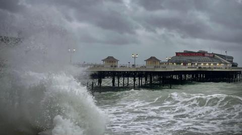 The pier at Cromer with a choppy murky sea below and a huge wave crashing against the wall, at the front of the photo. At the end of the pier is Pavilion Theatre.