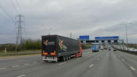 Three lanes of the M1 with two slip roads to the left, and blue motorway signs overhead. There is a line of lorries in the left-hand lane and cars in the other lanes.