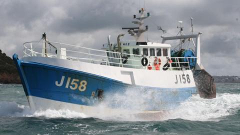 A white and blue fishing boat with J158 on the left side, in the ocean, next to another vessel out at sea with grey skies and land seen in the background.
