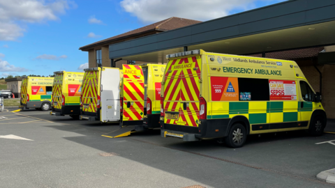 Five West Midlands Ambulance Service ambulances are parked in a row outside a building.