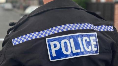 A close-up of the back of a police jacket. The word police is written in capital letters in white against a navy blue background in a rectangular box. A strip of blue and white chequered pattern is running between the two shoulders of the jacket.
