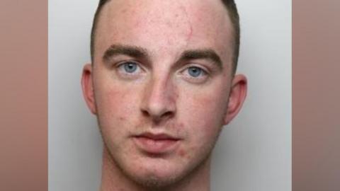 A police mug shot of a young man with short brown hair and blue eyes looking directly into the camera