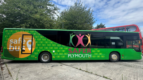 A view of the side of the bus. The bus is green and has stickers promoting Safer Plymouth