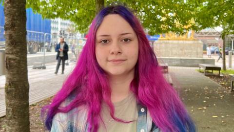 A woman with bright pink and purple hair looking into the camera. She is wearing a green top with a blue and white check over-shirt. Behind her is a number of trees in a pedestrian square that has benches to sit on. 