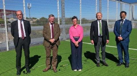 Four men and a woman stand side-by-side on a football pitch. The men wear suits and ties, and the woman wears a pink shirt and blue trousers.