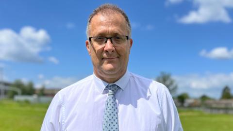 Vic Goddard stands on the school playing field in white shirt and blue tie