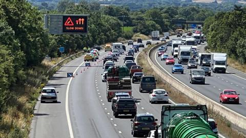 Traffic jam on the M5 motorway in Somerset. One lane is closed off by police. The gantry shows the lane closure and advises motorists to slow down.