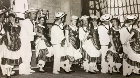 A black and white image showing men and women on stage. The men are in light colour sailor-type costumes and the women are wearing darker coloured dresses. Their costumes have a naval theme.