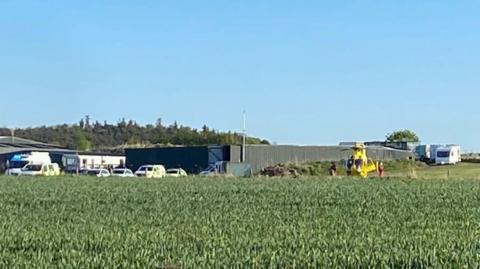 Several emergency service vehicles including an ambulance helicopter in the middle of a field. Campervans can be seen in the background