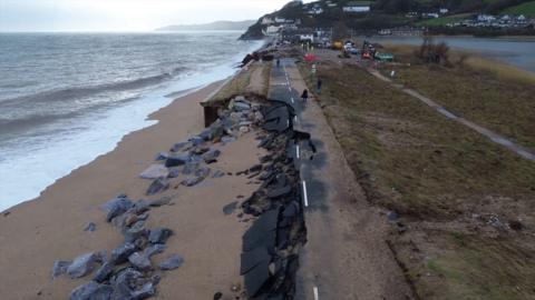 A picture of the washed away road. There is a number of rocks and sand covering the road next to the sea front.