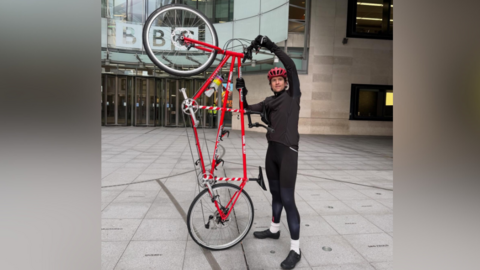 Greg James holding red tandem bike outside of the BBC's New Broadcasting House building in London