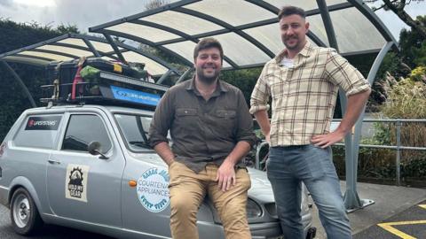 Two men in front of a Reliant Robin vehicle. The man on the left is wearing a chequered shirt and blue jeans, he is holding his hands on his hips. The man on the left is sitting on the bonnet of the vehicle. He is wearing beige chinos and a brown shirt. The car is silver and has a number of stickers on its chassis. There is a roof rack on the top of the car.
