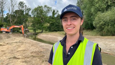 Sophie Bloor looks into the camera and is smiling. She is wearing a navy blue cap with 'Severn Rivers Trust' written on it, a navy blue polo shirt and a hi-vis vest. She is stood in front of a freshly-dug channel with water running through it. In the distance is a bright orange digger, and trees and bushes lining the edge of the field. 