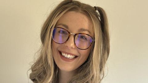 A smiling woman with long blonde hair. She is wearing glasses and posing in front of a cream wall. 
