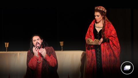 A photograph from the Macbeth play. There is a man kneeled on the ground wearing red. Then a woman is on the right holding a crown and wearing a crown in red robes. 