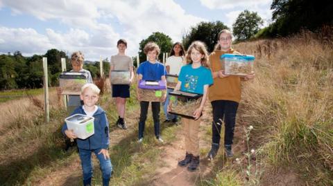 Seven children are smiling and holding clear boxes filled with straw and mice.