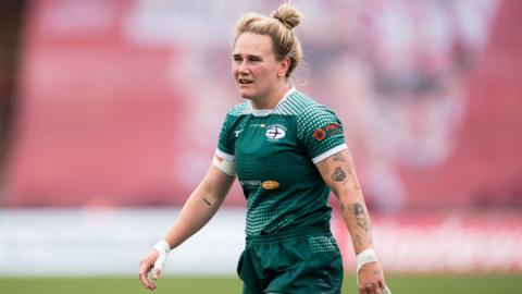 Meg Jones looks on, wearing the green home kit of Ealing Traifinders, showing a number of tattoos down her left arm.