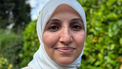 A close-up of a woman's face, looking at the camera. She wears a very light green hijab. She stands in front of some bushes, which are blurred.