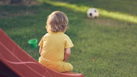 A young boy sits on the end of a slide in a play area, looking at a football