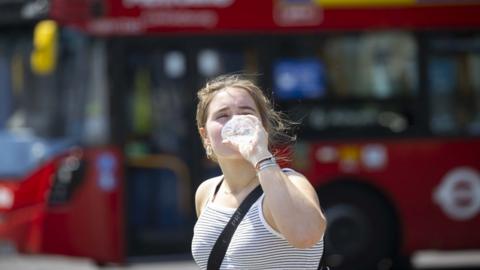 A young woman wearing a t shirt drinks from a bottle of water as she stands on a pavement with a red bus driving past behind