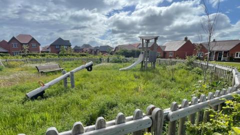 An overgrown playground with long grass is pictured on a sunny day. There are houses all around the site with a seesaw, a slide and a bench in the shot. 