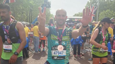 Chris at the finish line of the London Marathon, wearing his medal. His hands are aloft, one in a peace sign. Other finishers are around him.