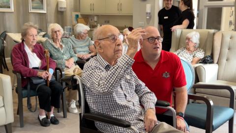 An elderly man in a wheelchair about to throw a dart at a darts board in the lounge of Heatherton House care home.