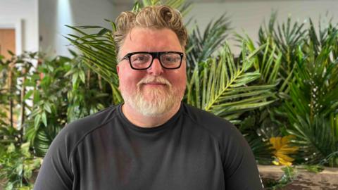 A man with a beard and wearing black-rimmed glasses stands in front of some green houseplants inside while being interviewed 