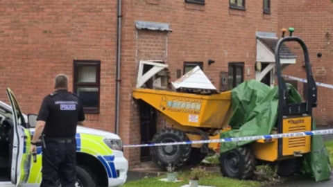 A yellow mini digger is pushed up against the front of a house, destroying the canopy over the door and brickwork surrounding the entrance. Police tape surrounds the house, and a policeman standing by his patrol car looks on.