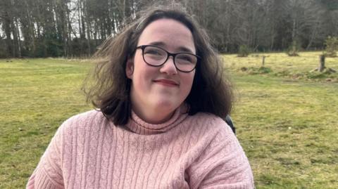 Iona Grant has dark hair down to her shoulders and is wearing a hair band and glasses. She is also wearing a pink woolly jumper. She is in a wheelchair in the outdoors. There is grass and woodland behind her.