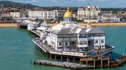 An aerial view of Eastbourne Pier shows a large white pavilion building at the end featuring a central gold dome and smaller domes. The structure stands on dark metal supports above the sea, with wooden walkways and railings visible around its perimeter. Behind the pier, seafront hotels and apartments line a sandy beach, with hills in the background.