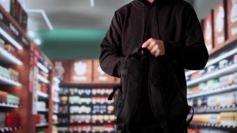 A person wearing a hooded sweatshirt stands in a supermarket aisle, holding a backpack, with shelves of packaged food products on both sides.