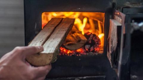 A man places a log on a wood burning stove. The fire in the stove is alight and the door is open. Flames, embers and burning wood are in the stove.
