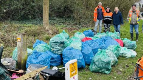 Three people, standing by a large amount of rubbish, including blue and green bin bags. 