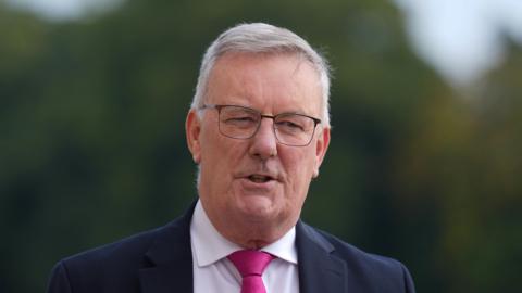 Mike Nesbitt stares into the camera, his hair is grey and his mouth is slightly open. He has on a white shirt, pink tie and black suit. Behind him green tree's are blurred.