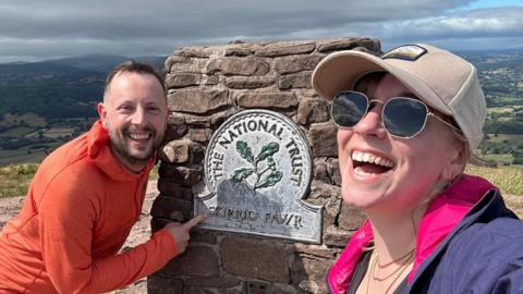 Jody and Jessie stand on Skirrid Fawr walk. Jody is wearing an orange half zip jacket. Jessie is taking a selfie and wearing a purple jacket, white hat and sunglasses. 